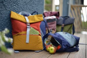 One rucksack propped up against a wall in orange and yellow, one rucksack on it's side with apples spilling out of it, in blue and grey.
