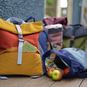One rucksack propped up against a wall in orange and yellow, one rucksack on it's side with apples spilling out of it, in blue and grey.